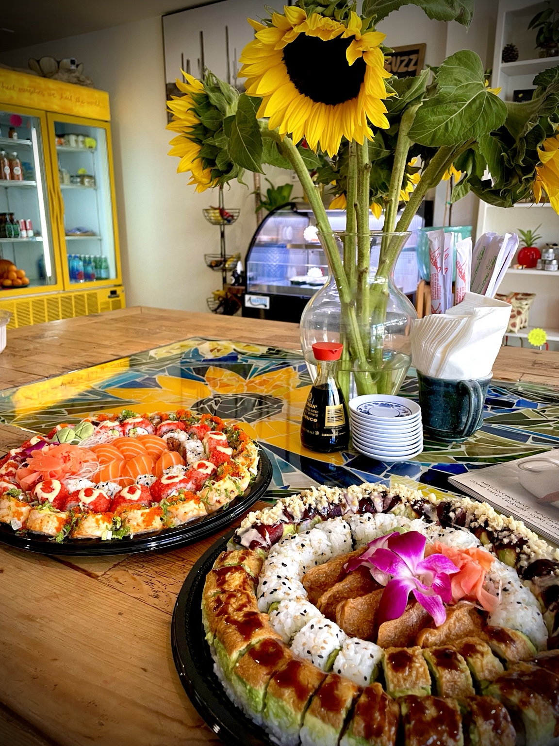 Sushi platters at the counter beside a vase of sunflowers